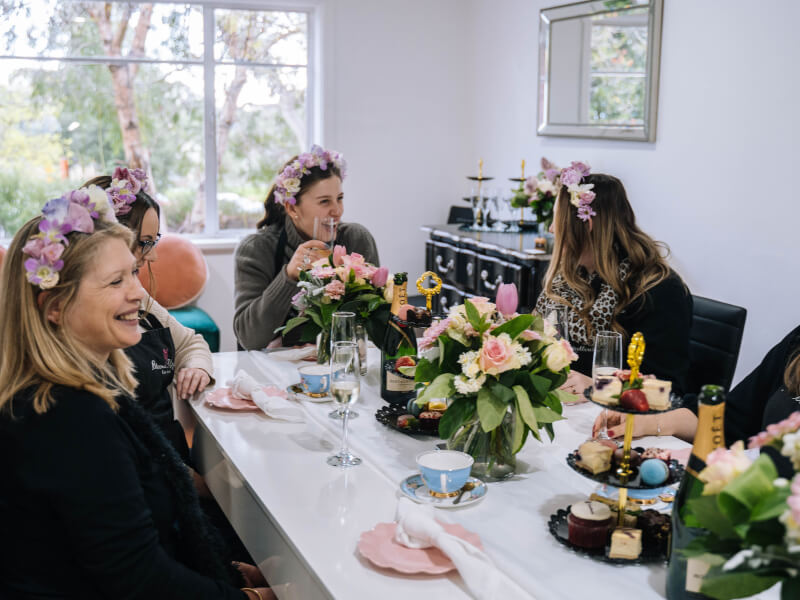 A group of women wearing flower crowns sit at a table enjoying a hen do.